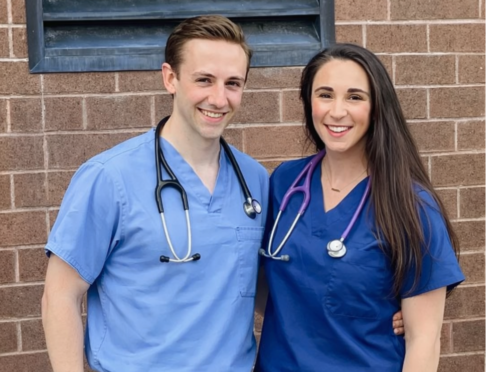 Two individuals in blue medical scrubs standing against a brick wall. 